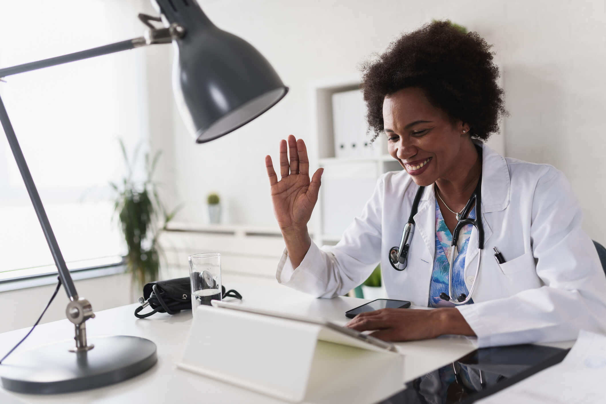 Médica de pele negra sorrindo sentada em mesa olhando para tablet com a mão levantada