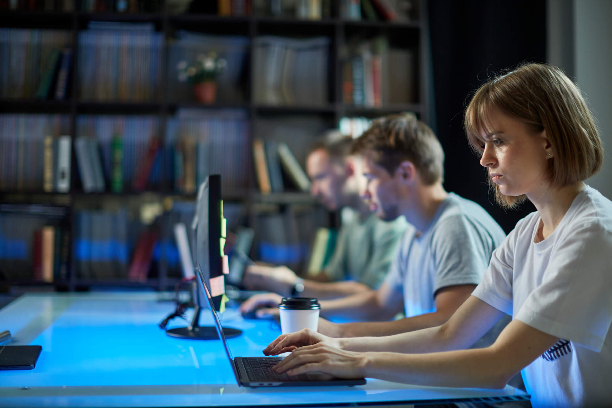 uma mulher e dois homens sentados lado a lado em uma mesa digitando em notebooks e computadores em um ambiente com estante de livros ao fundo