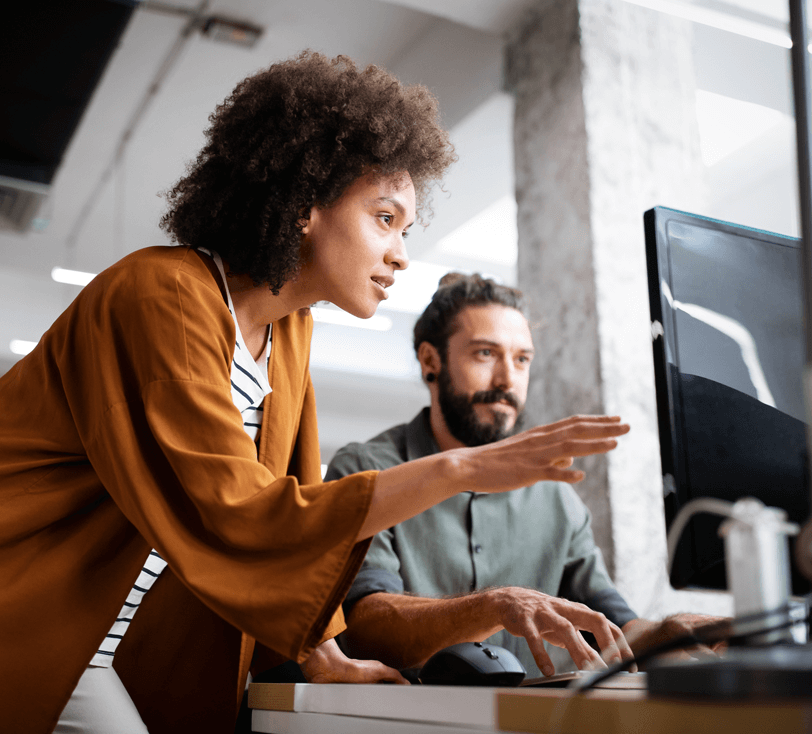 Mulher negra de cabelos crespos olhando e apontado para computador enquanto homem branco de barba olha na mesma direção